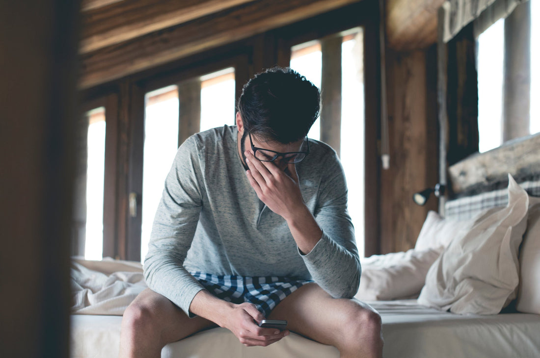 Tired young man with a smartphone sitting on the bed at home squinting.