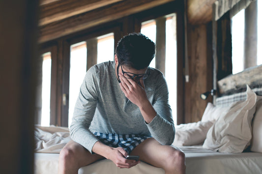 Tired young man with a smartphone sitting on the bed at home squinting.